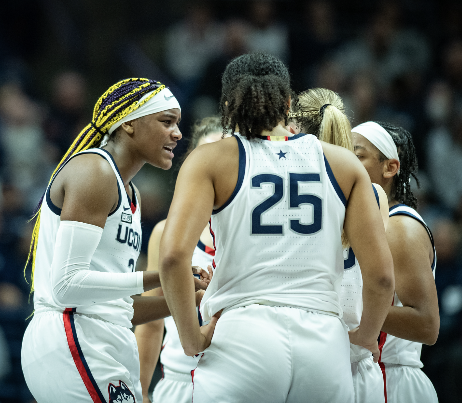 Members of the UConn women's basketball team gather in a huddle. They are wearing home white jerseys. The back of Ice Brady's No. 25 jersey is pictured in the forefront.